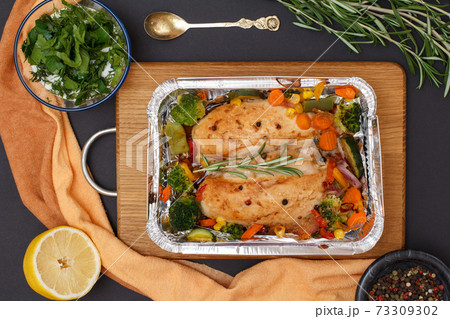 Baked chicken breasts or fillet in metal container with vegetables on cutting board. Baked chicken breasts or fillet in metal container with vegetables on cutting board. 73309302