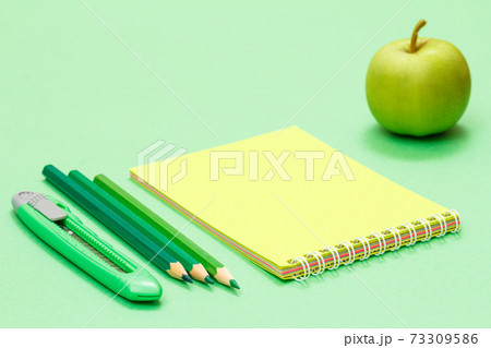 School supplies. Paper knife, color pencils, notebook and apple on the green background. School supplies. Paper knife, color pencils, notebook and apple on the green background. 73309586