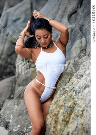 Beautiful girl in a white bikini posing on a beach with rocks Beautiful girl in a white bikini posing on a beach with rocks 73310936