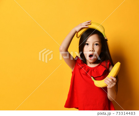 Asian female in red blouse, multicolored skirt. She looking wondered, holding bananas, posing on orange background. Close up Asian female in red blouse, multicolored skirt. She looking wondered, holding bananas, posing on orange background. Close up 73314483