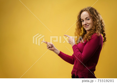 Portrait of young woman in a studio on yellow background, pointing fingers to copy space. 73319201