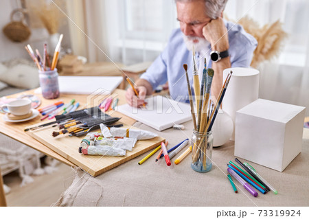 paintbrushes in glass cans and gouache, paints, pencils on table and artist in the background 73319924