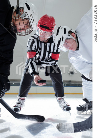 Hockey referee holding puck over ice rink while rivals with sticks looking at it 73322026