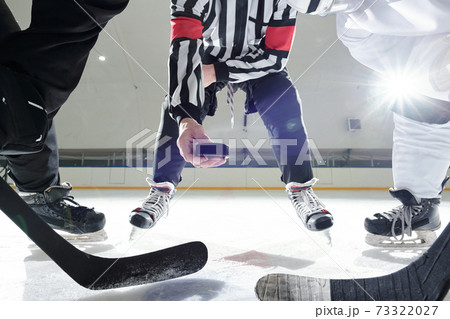 Hockey referee with puck standing on ice rink between two players with sticks 73322027
