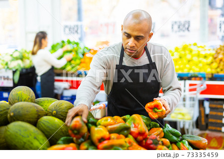 Merchandiser in an apron lays out ripe bell peppers on supermarket counter Merchandiser in an apron lays out ripe bell peppers on supermarket counter 73335459