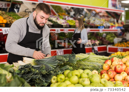 Male shop assistant in apron lays fresh leek in supermarket Male shop assistant in apron lays fresh leek in supermarket 73340542