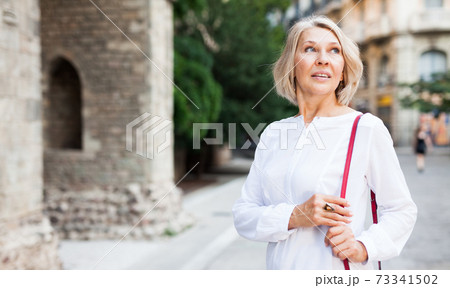 Cheerful mature female is walking in white blouse on the street outdoor Cheerful mature female is walking in white blouse on the street outdoor 73341502