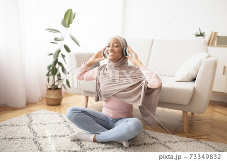 Full length portrait of black Muslim woman in hijab and headphones listening to music on floor in living room Full length portrait of black Muslim woman in hijab and headphones listening to music on floor in living room 73349832