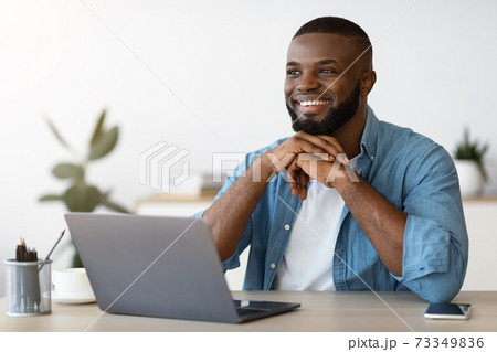 Portrait of pensive smiling black freelancer sitting at desk in home office Portrait of pensive smiling black freelancer sitting at desk in home office 73349836