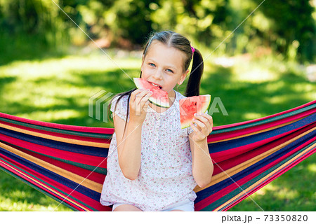 Little girl eating watermelon slices in the garden sitting on a hammock Little girl eating watermelon slices in the garden sitting on a hammock 73350820