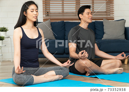 Healthy young Asian couple attractive couple practicing yoga exercises sitting on a mat in lotus pose in an online class on quarantine at home. Concept of Technology for New normal lifestyle 73352938