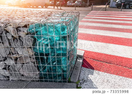Close-up detail of new modern beautiful gabion fence with metal cage filled by crushed stone and shattered artificial blue glass rocks near zebra crossroad. City street road pavement protection 73355998