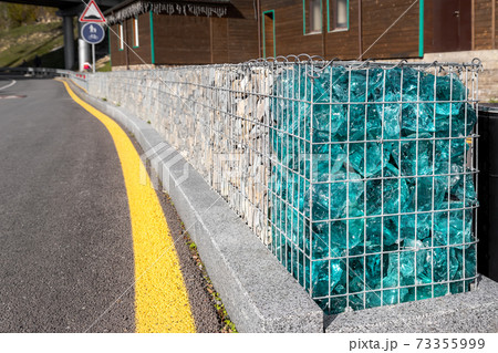 Close-up detail of new modern beautiful gabion fence with metal cage filled by crushed stone and shattered artificial blue glass rocks near asphalt roadway. City street road pavement protection 73355999