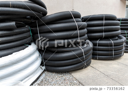 Many rolled colis of new black corrugated goffred plastic electric plumbing drainage pipe prepared for installation near building construction site. Drain sewage tube hose conduit pipeline 73356162