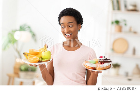 Cheerful black woman holding plates with fruits and desserts indoors, choosing between healthy and junk foods Cheerful black woman holding plates with fruits and desserts indoors, choosing between healthy and junk foods 73358087