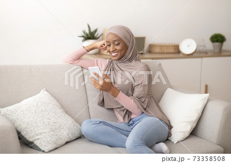 Millennial Muslim lady in hijab using mobile phone at home, messaging or browsing social media, sitting on couch indoors 73358508