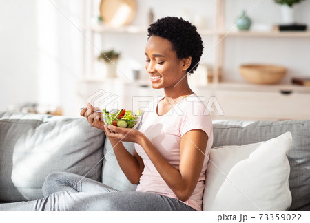 Balanced nutrition concept. Lovely black lady eating delicious vegetable salad on cozy couch at home Balanced nutrition concept. Lovely black lady eating delicious vegetable salad on cozy couch at home 73359022
