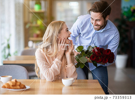 Celebrating special occasion. Romantic young guy presenting bouquet of red roses to his excited girlfriend at cafe Celebrating special occasion. Romantic young guy presenting bouquet of red roses to his excited girlfriend at cafe 73359313