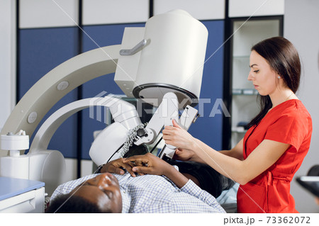 Close up side view of high-skilled female doctor in red uniform, performing lithotripsy, using modern lithotripter to break up kidney stones of male African patient with high-intensity acoustic pulse Close up side view of high-skilled female doctor in red uniform, performing lithotripsy, using modern lithotripter to break up kidney stones of male African patient with high-intensity acoustic pulse 73362072