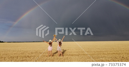 In a wheat field after the rain, two girls run. Colorful real rainbow over the horizon. europe Ukraine. Summer crop fields, ripening wheat field. 73375374