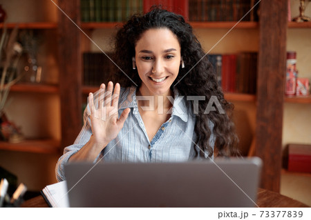Woman sitting at desk, using laptop and waving to webcam 73377839