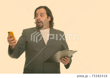 Fat Caucasian businessman looking shocked while using mobile phone and holding clipboard Fat Caucasian businessman looking shocked while using mobile phone and holding clipboard 73382419