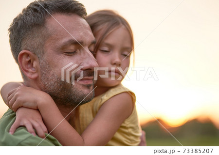 Close up shot of young father and adorable little daughter embracing each other, posing with eyes closed while spending time outdoors in the park on a summer day 73385207