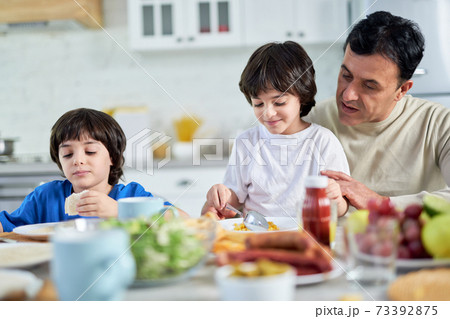 Loving middle aged hispanic father serving his cute little son, sitting with kids at kitchen table while having a breakfast at home Loving middle aged hispanic father serving his cute little son, sitting with kids at kitchen table while having a breakfast at home 73392875