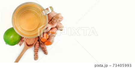 ginger and turmeric tea in glass cup next to lemon, cinnamon wooden spoon on white background with copy space	 73405993