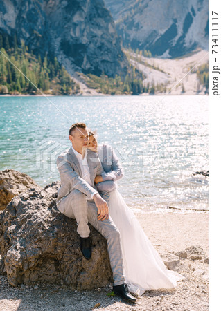 Bride and groom sit embracing against background of stones at the Lago di Braies in Italy. Destination wedding in Europe, on Braies lake. Loving newlyweds walk against the backdrop of amazing nature Bride and groom sit embracing against background of stones at the Lago di Braies in Italy. Destination wedding in Europe, on Braies lake. Loving newlyweds walk against the backdrop of amazing nature 73411117