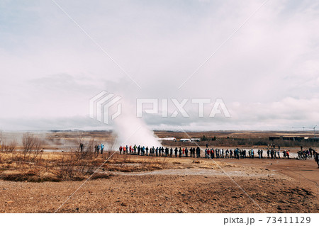 Tourists look at the eruption of a geyser. Geyser Valley in southwest of Iceland. The famous tourist attraction Geysir. Geothermal zone Haukadalur. Strokkur geyser on the slopes of Laugarfjall hill. 73411129