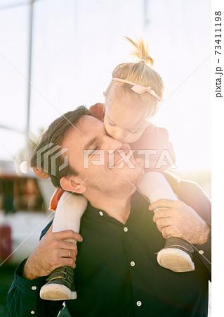 2-year old is sitting on her father's shoulders and kissing him on the cheek 73411198
