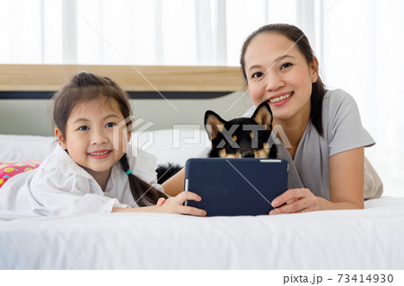 Happy Asian mother and daughter lying on bed and using digital tablet. Black Shiba Inu on bed between its owner. Concept family with dog at home. 73414930