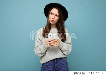 Beautiful young brunette woman thinking wearing black hat and grey sweater holding smartphone 73415407