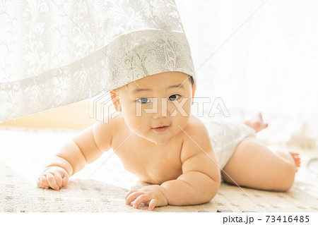 窓辺ではいはいをしながら見つめてくる裸の赤ちゃん 73416485
