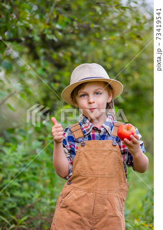 Portrait of one cute boy in a hat in the garden with a red apple, emotions, happiness, food. Autumn harvest of apples. 73419541