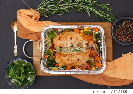 Baked chicken breasts or fillet in metal container with vegetables on cutting board and kitchen napkin. 73423805