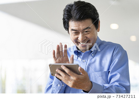 スマートフォンの画面に向かって手を振る笑顔のシニア男性 73427211