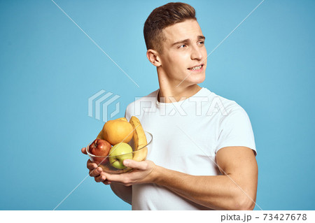 Happy man with fresh fruits gesturing with hands blue background white t-shirt vitamins bananas oranges apples 73427678