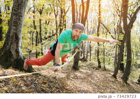 A bearded man in age balances while sitting on a taut slackline in the autumn forest. Outdoor Leisure 73429723