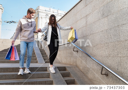 Tourists walk around city with purchases in their hands. Guy helps girl to go down the stairs 73437303