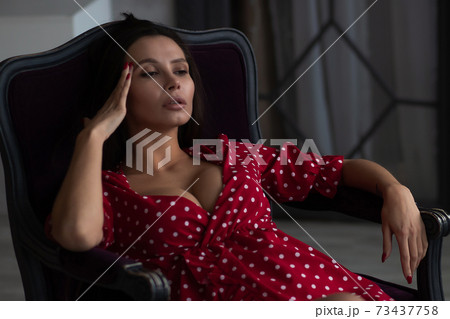 Alluring young woman in feminine dress resting in armchair 73437758