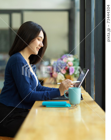 Female student doing assignment with digital tablet and stationery in cafe 73444304