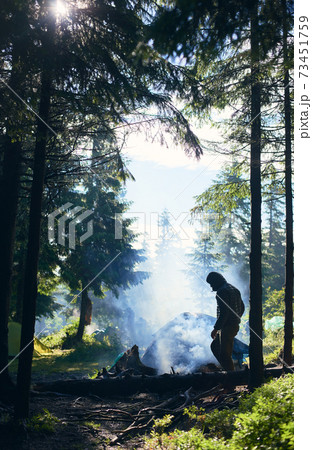 Tent among fir trees under the rays of sun breaking through clouds on cold day. Guy making fire and smoke flows up 73451759