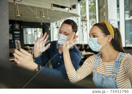 Two people wearing face masks, using a smart phone, waving during a face time call. 73452790