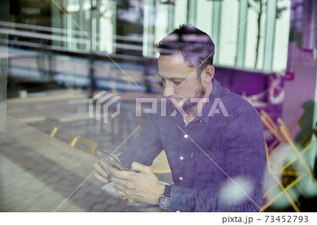 Man in a face mask seated at a cafe table using a mobile phone, view through a window 73452793
