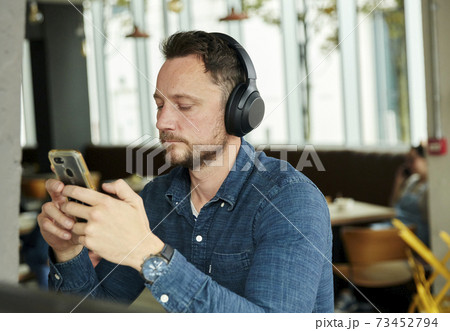Man seated in a cafe wearing headphones, using a smart phone, working remotely. 73452794
