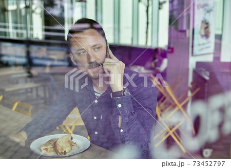Man in a face mask seated at a cafe table using a mobile phone, view through a window 73452797