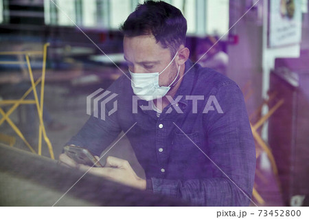 Man in a face mask seated at a cafe table using a mobile phone, view through a window 73452800