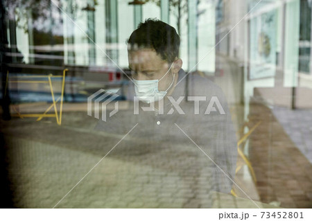 Man in a face mask seated at a cafe table, view through a window 73452801